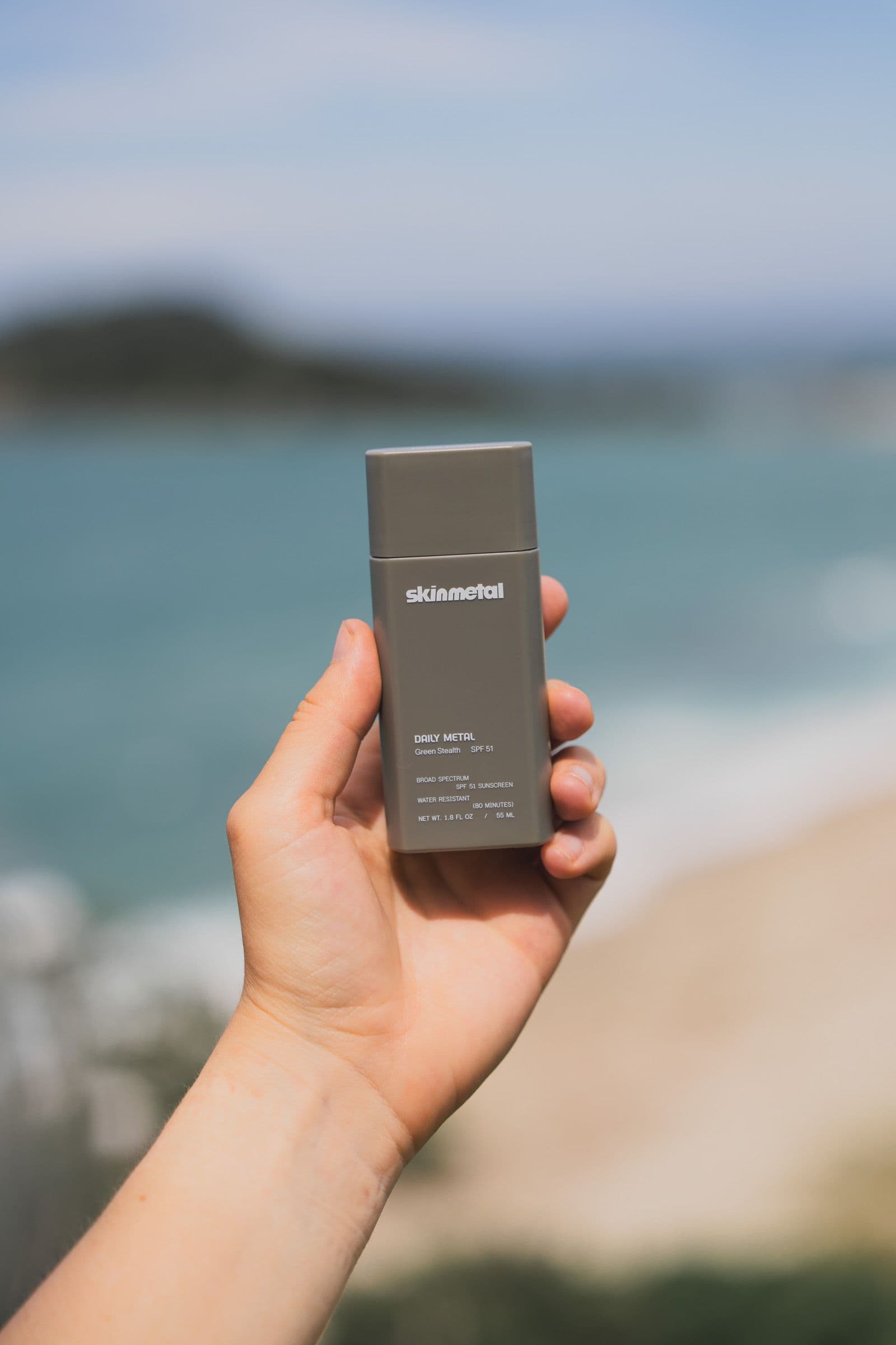 A hand holds a gray Skinmetal sunscreen bottle over a blurred teal bay and beach.