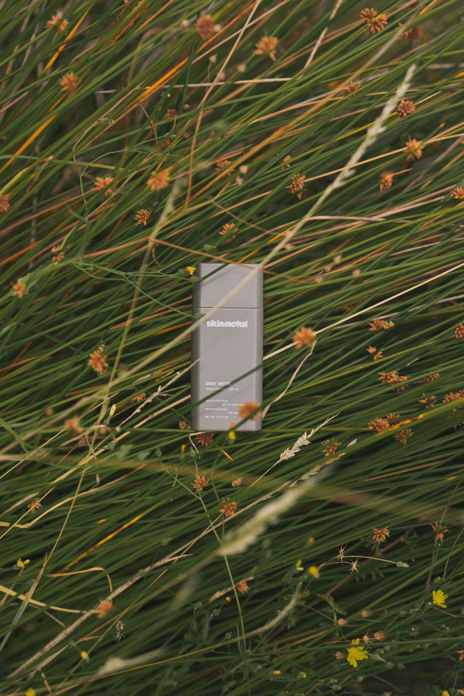 A gray Skinmetal sunscreen bottle rests among green grass and orange seed heads.