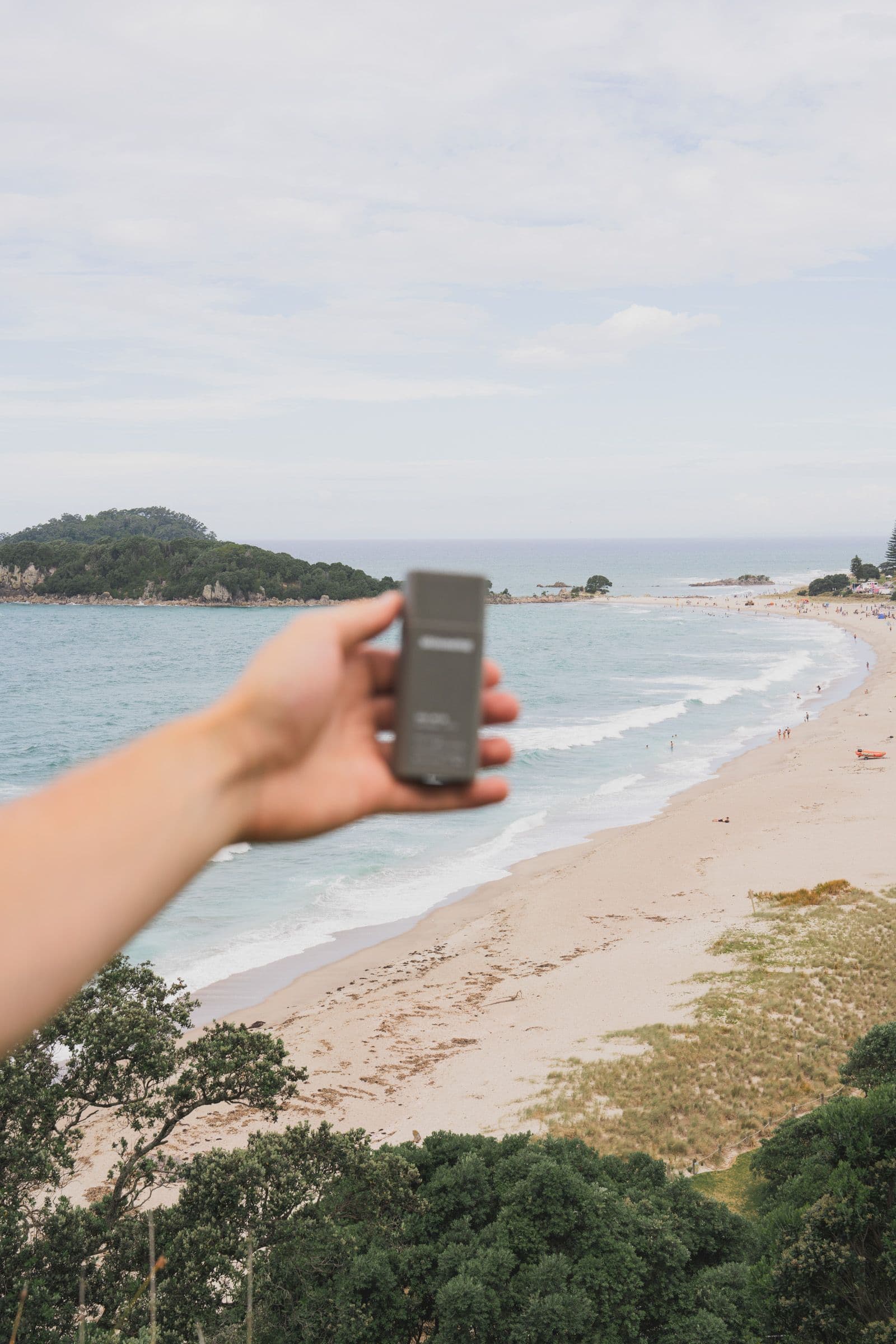 A hand holds a small gray product over a wide beach and calm ocean horizon.