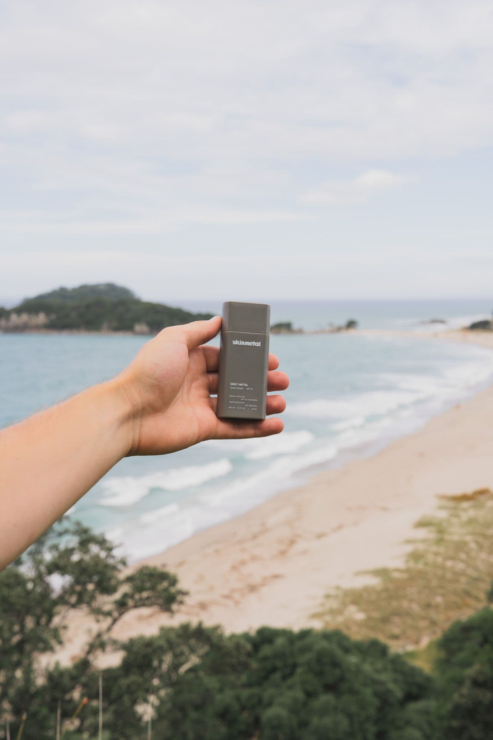A hand holds a gray Skinmetal sunscreen above a long sand beach with breaking waves.