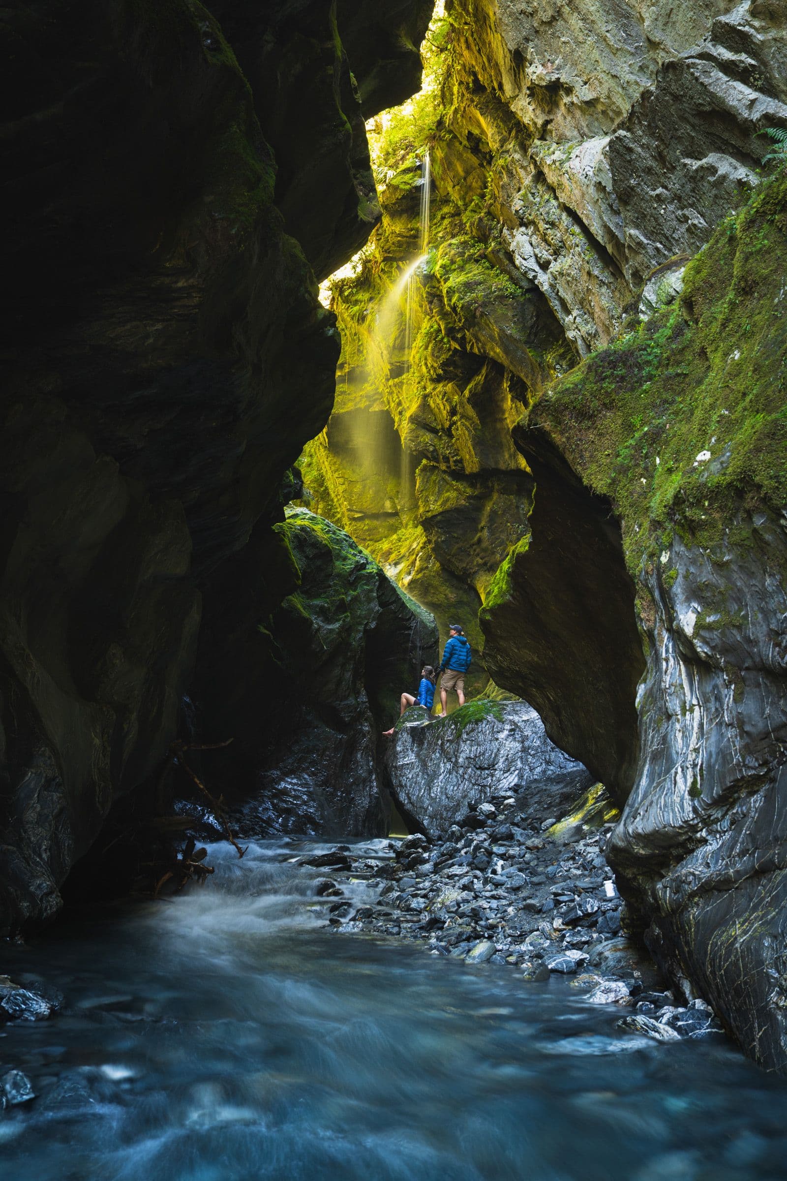 Two hikers stand on rocks inside a narrow mossy canyon with a thin waterfall trickling down and a stream flowing below.