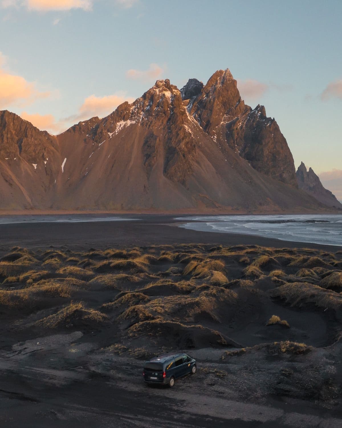 Black sand and tufts of grass below Vestrahorn mountain at golden hour, small camper parked nearby
