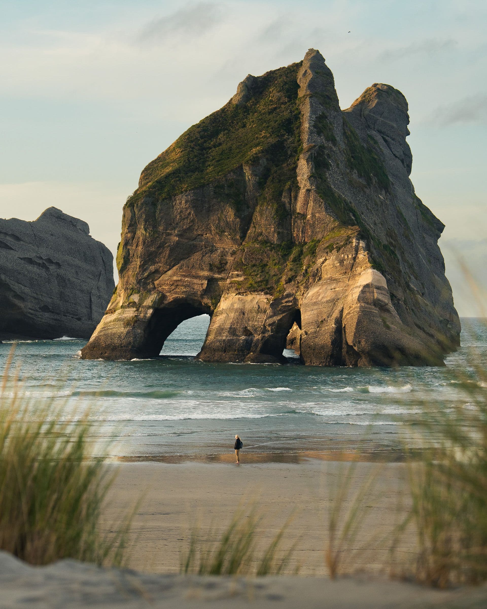 Tall rocky sea arch on sandy beach with lone person standing small at the water's edge.