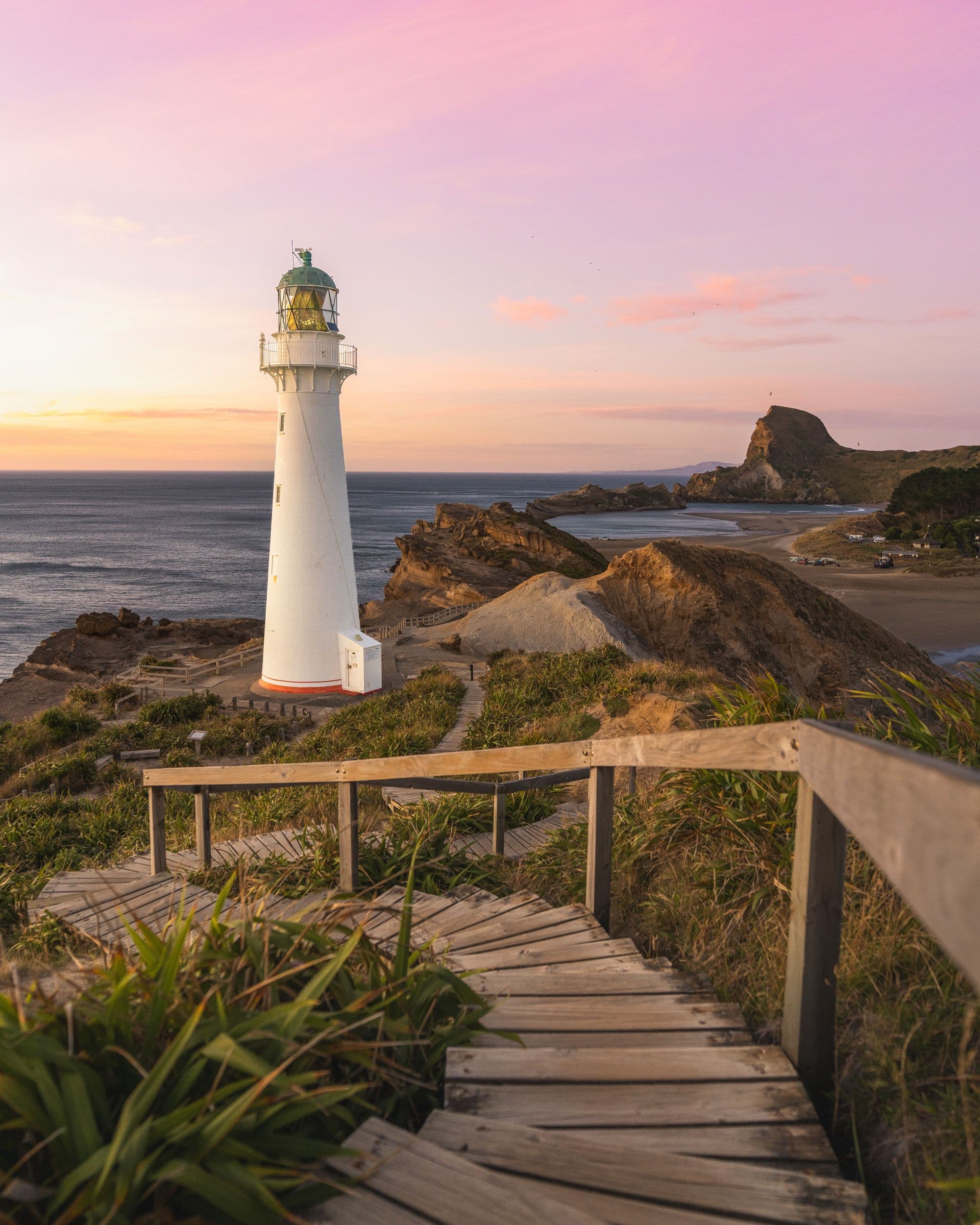 White lighthouse on a rocky headland above the sea as a pink and peach sunset fills the sky.