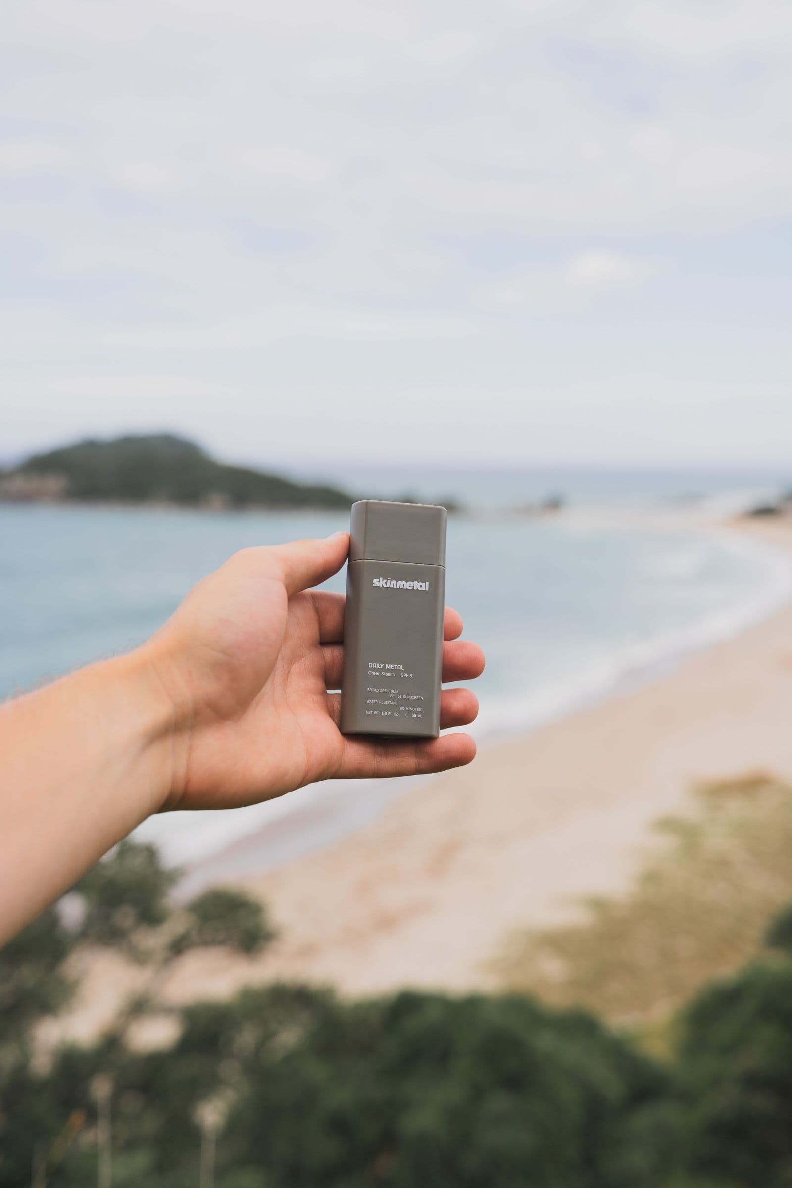 A hand holds a gray Skinmetal sunscreen above a soft-focus ocean bay and beach.