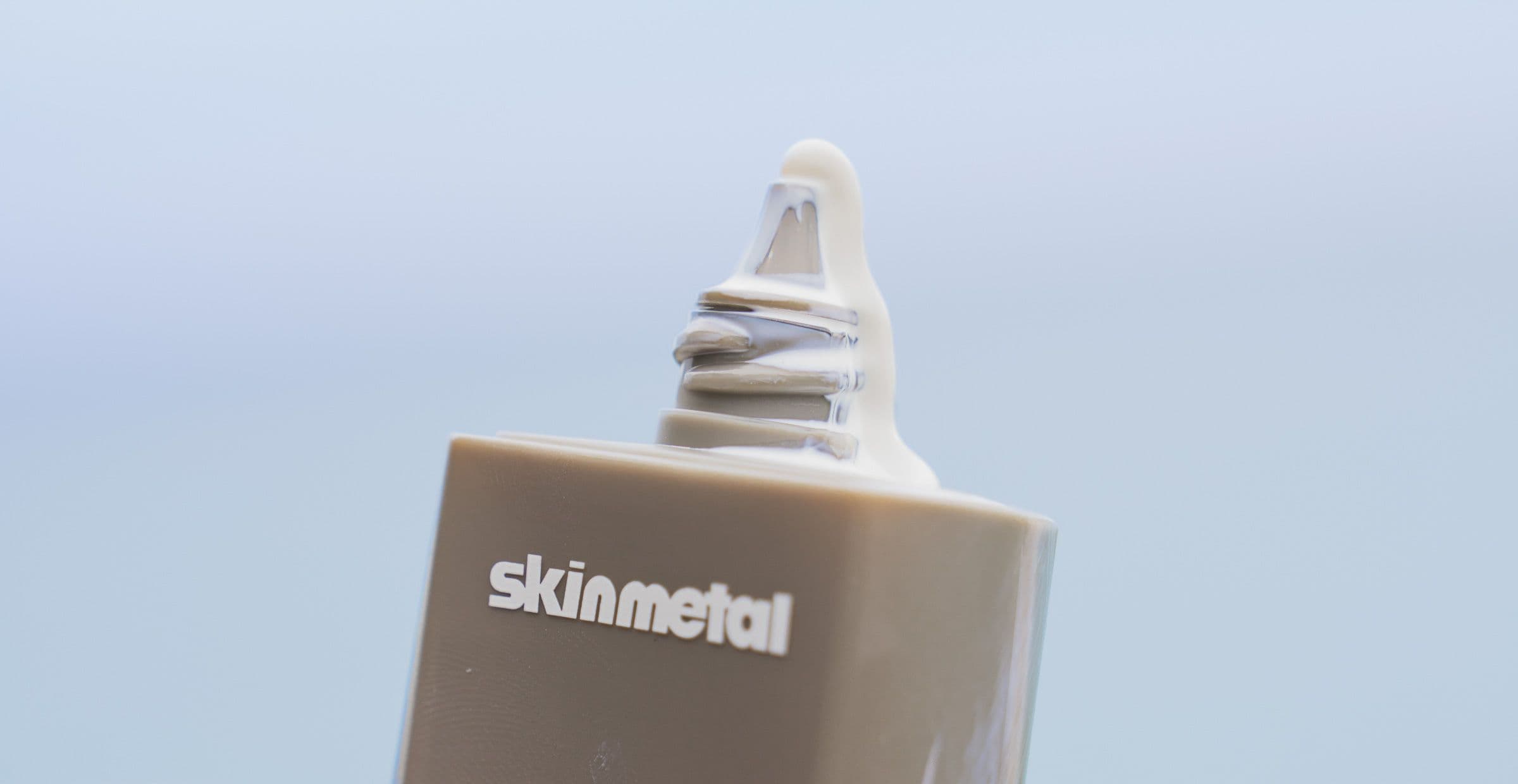 Close-up of a Skinmetal sunscreen nozzle with a drip of cream against soft blue sky.