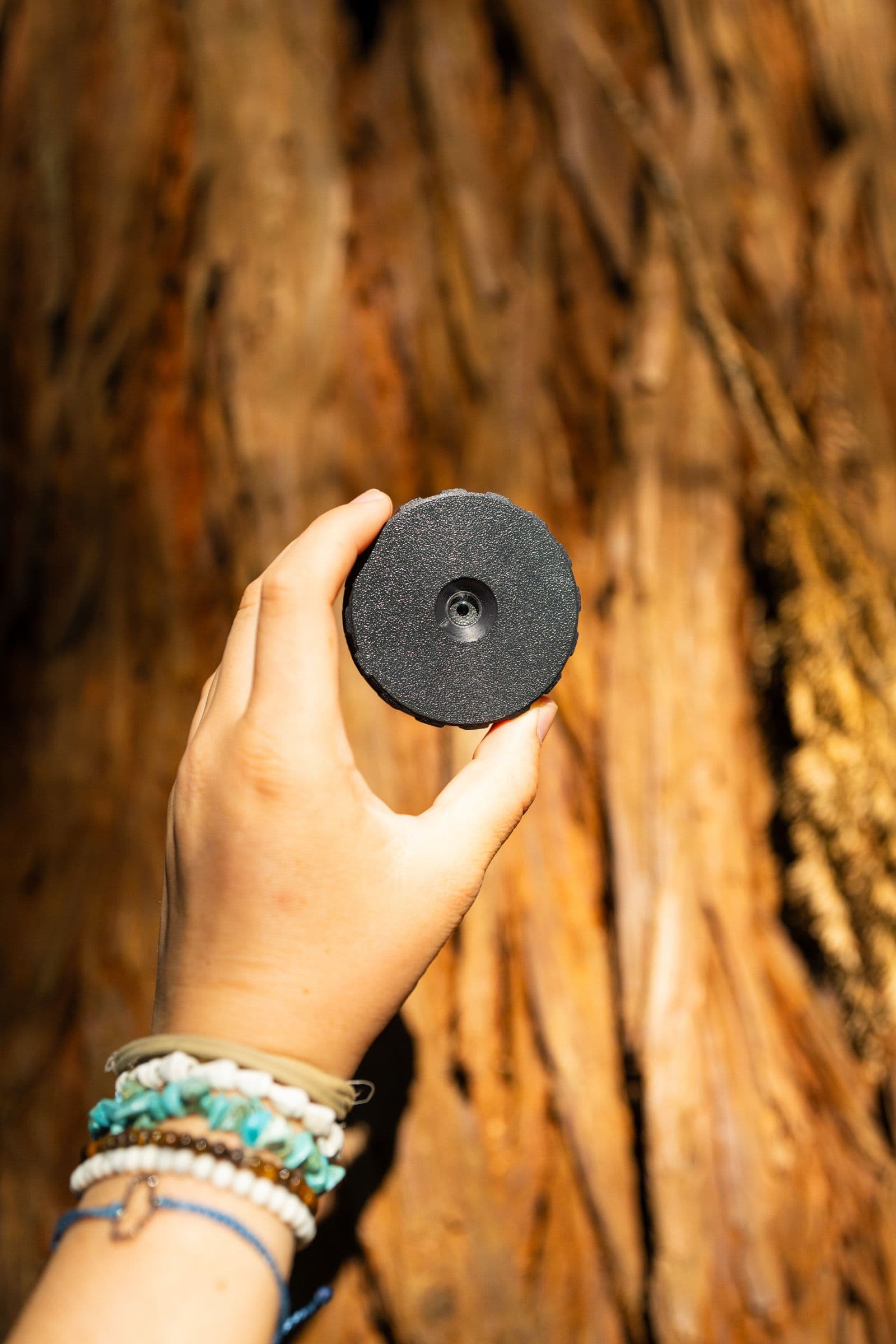 A hand with beaded bracelets holds a round black product against redwood bark.