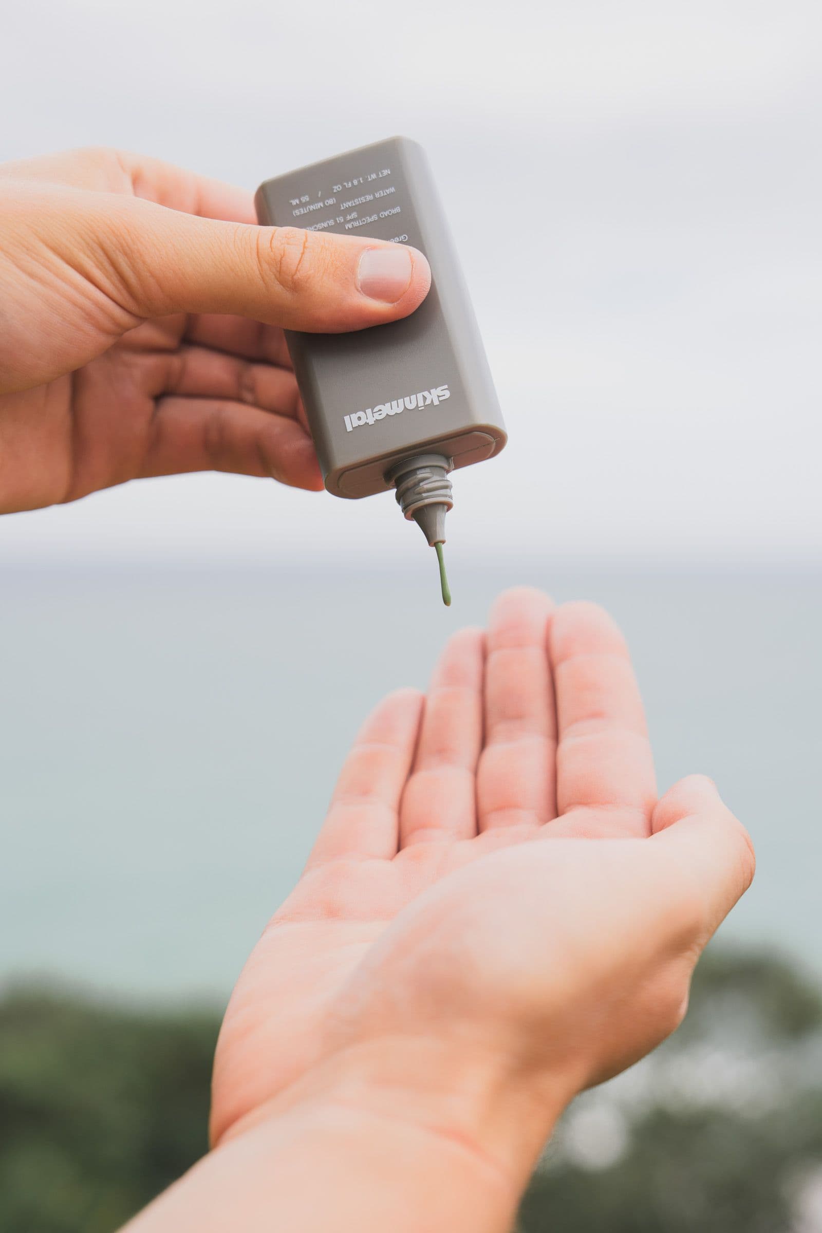 Green sunscreen streams from a Skinmetal bottle into an open palm above the ocean.
