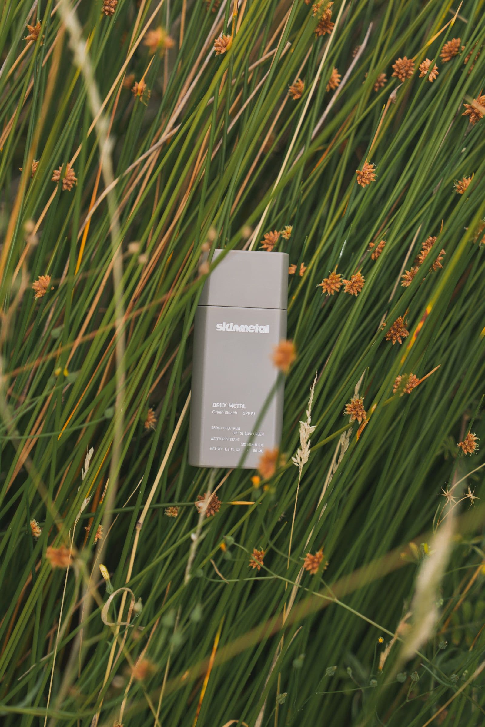 A gray Skinmetal sunscreen sits upright in a bed of green grass and small flowers.