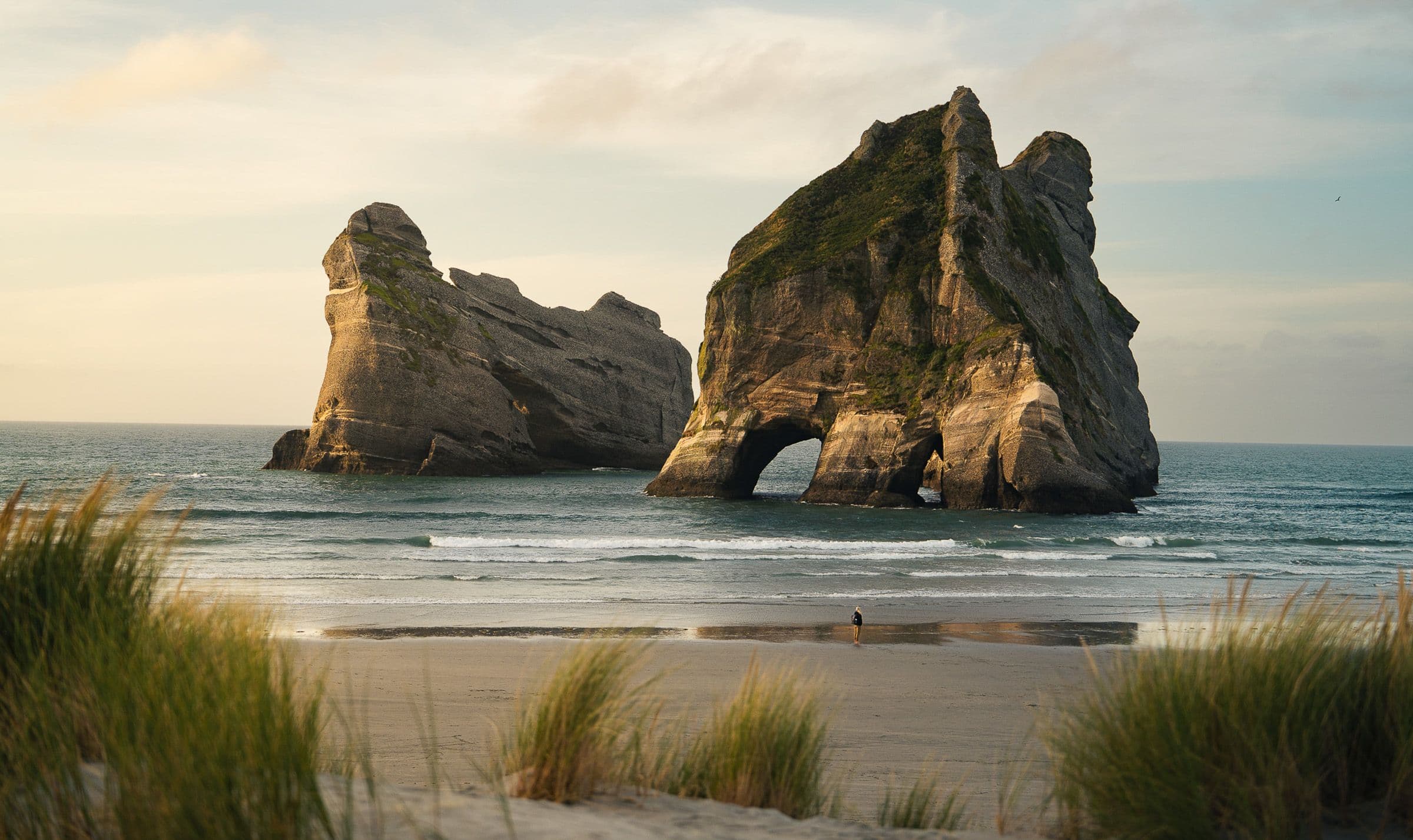 Large sea arch and rocky islands rise from ocean off sandy beach with dune grass in foreground.