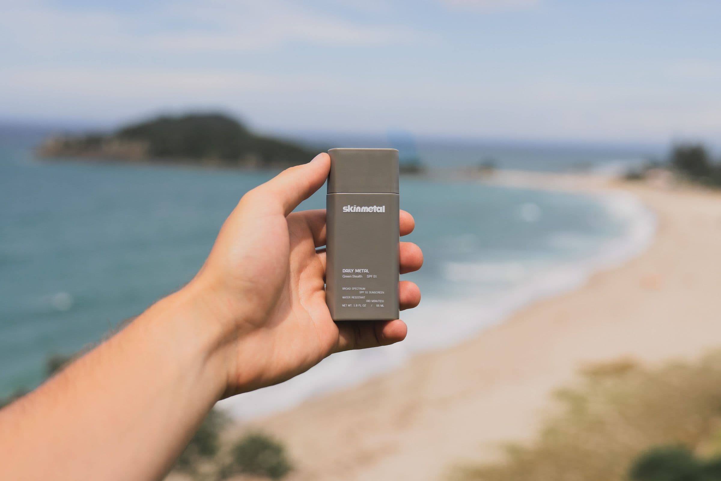 A gray Skinmetal sunscreen is held above a curving sand beach and blue ocean.