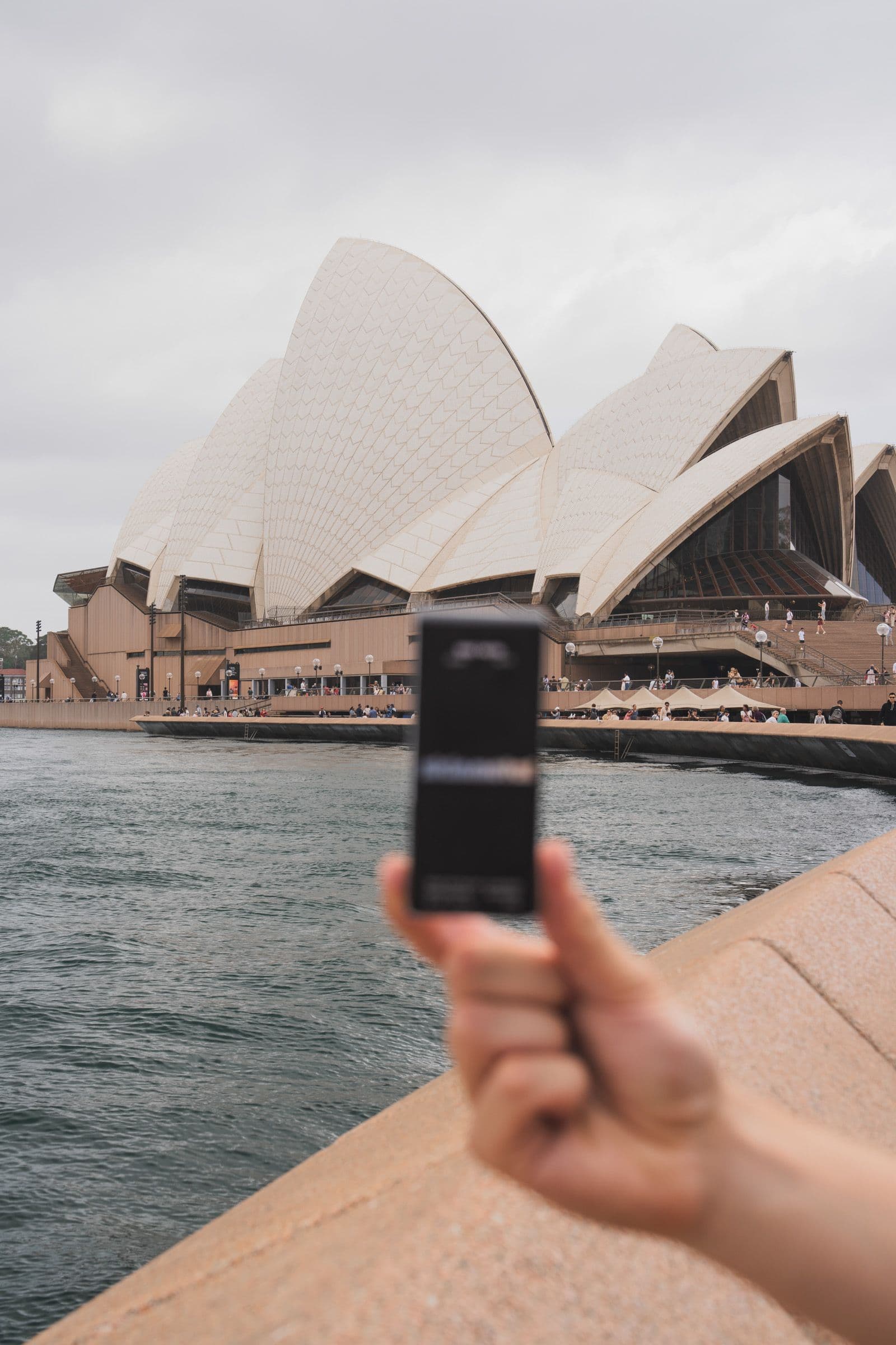 Hand holds small black product box in front of Sydney Opera House across harbor water.